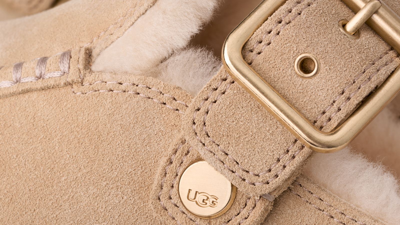 UGG Goldenstar Evelina Clog Sand Castle yakın plan: altın tonlu toka, UGG logolu düğme, mokasen dikişi ve koyun postu